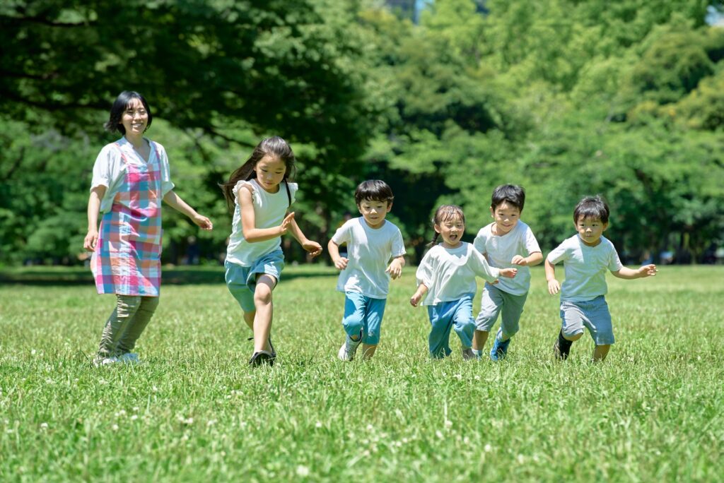 公園の芝生の上で、笑顔でかけっこを楽しんでいる保育士と子どもたち