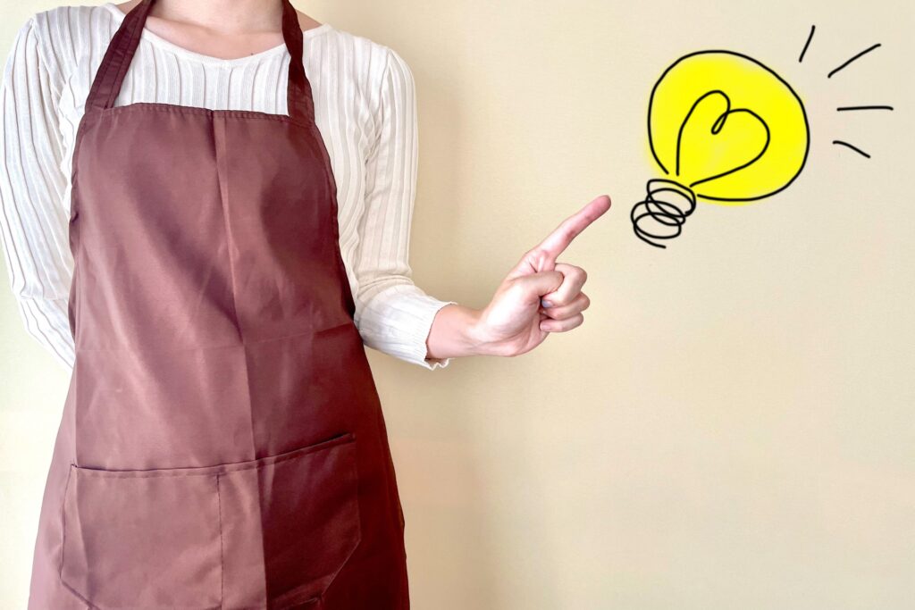 顔は見えていない茶色のエプロン姿の女性か人差し指を立ててひらめいている